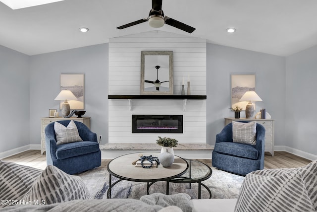 living area featuring ceiling fan, light wood-style flooring, a large fireplace, baseboards, and vaulted ceiling