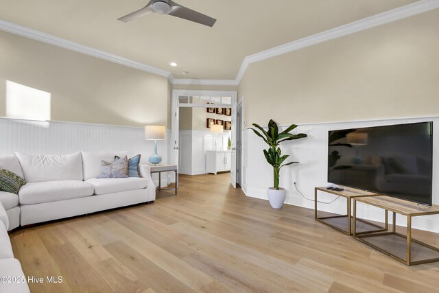 living room featuring light wood-type flooring, a wainscoted wall, a ceiling fan, and crown molding