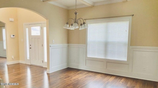 unfurnished dining area featuring arched walkways, wainscoting, wood finished floors, beamed ceiling, and a decorative wall