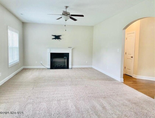 unfurnished living room with arched walkways, carpet floors, a fireplace with flush hearth, a ceiling fan, and baseboards