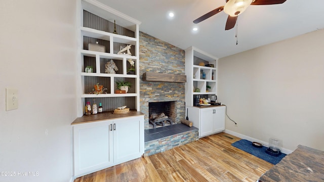 unfurnished living room with a stone fireplace, a ceiling fan, baseboards, built in features, and light wood-type flooring