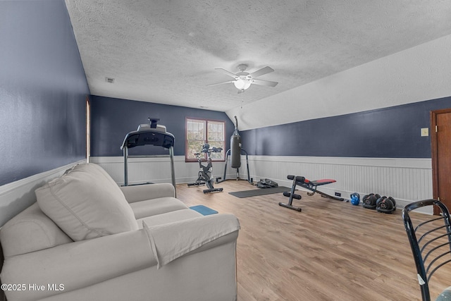 workout room with a textured ceiling, lofted ceiling, wood finished floors, a ceiling fan, and wainscoting