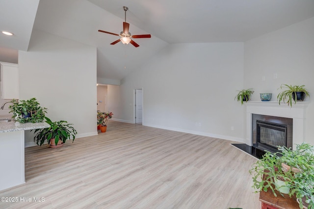 living area with baseboards, ceiling fan, light wood-style floors, a fireplace, and high vaulted ceiling