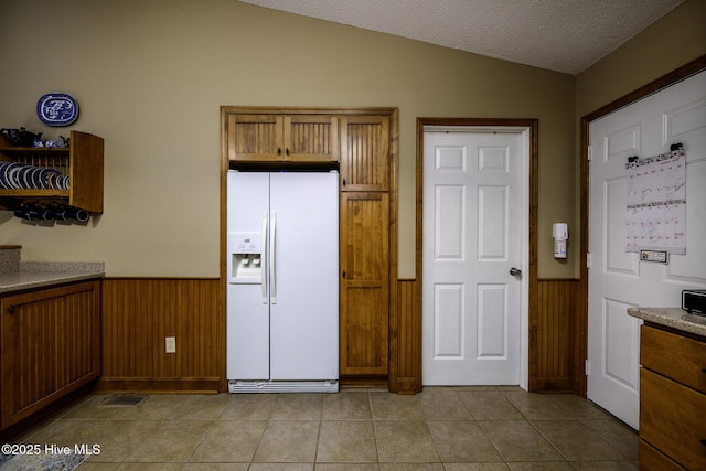 kitchen with a wainscoted wall, lofted ceiling, light tile patterned floors, brown cabinets, and white refrigerator with ice dispenser