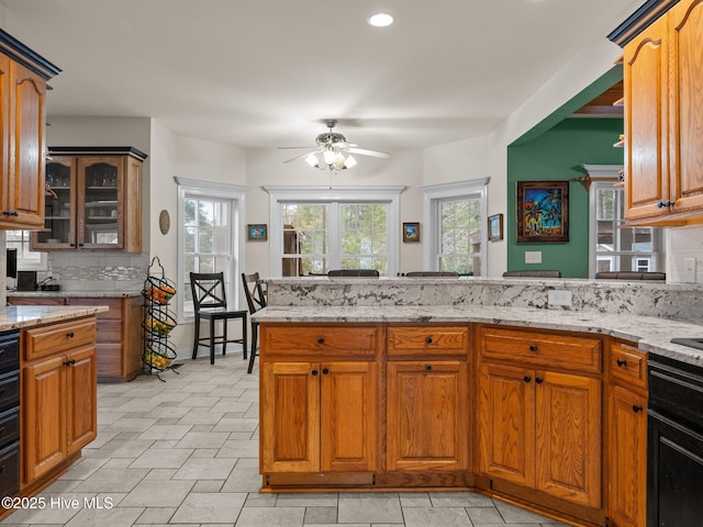 kitchen featuring tasteful backsplash, light stone counters, a peninsula, brown cabinetry, and a ceiling fan