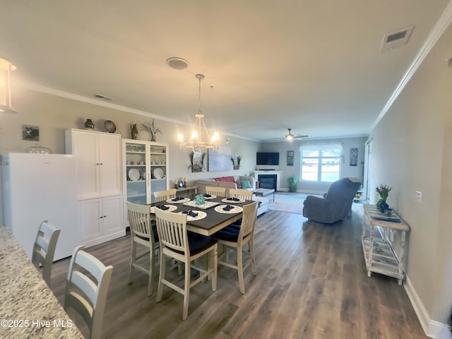 dining space featuring ceiling fan with notable chandelier, a fireplace, visible vents, baseboards, and dark wood-style floors