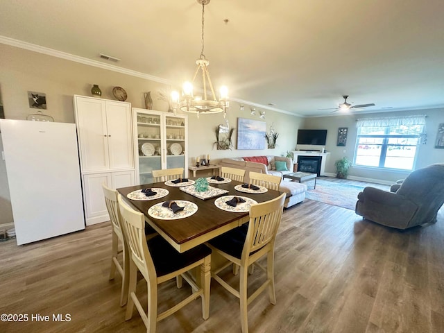 dining room with visible vents, ornamental molding, a glass covered fireplace, wood finished floors, and ceiling fan with notable chandelier