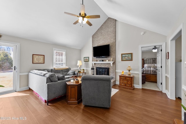 living room with baseboards, a ceiling fan, light wood-type flooring, a brick fireplace, and high vaulted ceiling