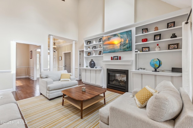 living room featuring built in features, wood finished floors, a high ceiling, decorative columns, and a glass covered fireplace