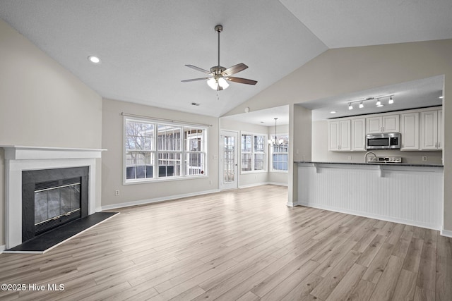 unfurnished living room with light wood-style flooring, ceiling fan with notable chandelier, baseboards, vaulted ceiling, and a glass covered fireplace