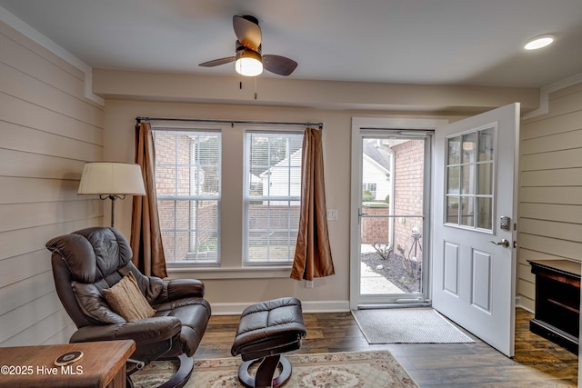 living area featuring a ceiling fan, wooden walls, baseboards, and wood finished floors