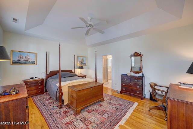 bedroom featuring a tray ceiling, light wood finished floors, visible vents, a ceiling fan, and ensuite bath