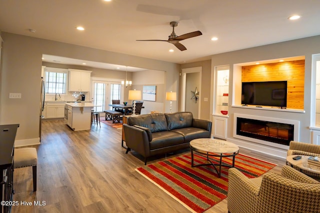 living area featuring baseboards, a glass covered fireplace, light wood-style flooring, ceiling fan with notable chandelier, and recessed lighting