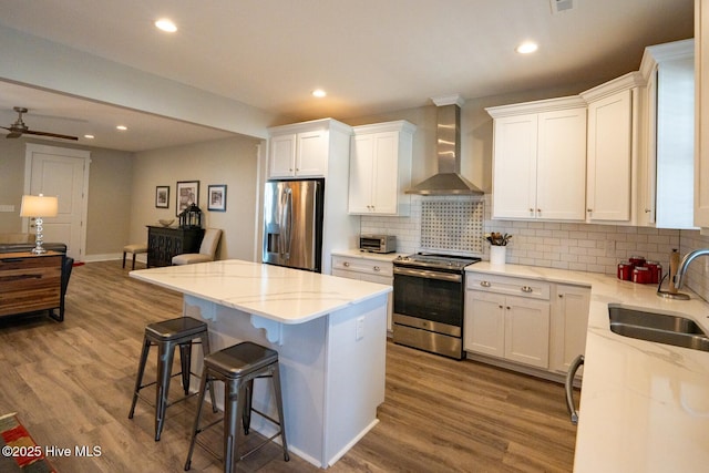 kitchen with a breakfast bar area, stainless steel appliances, wood finished floors, a sink, and wall chimney range hood