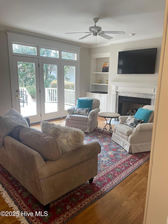 living area featuring built in shelves, wood finished floors, a premium fireplace, ceiling fan, and crown molding