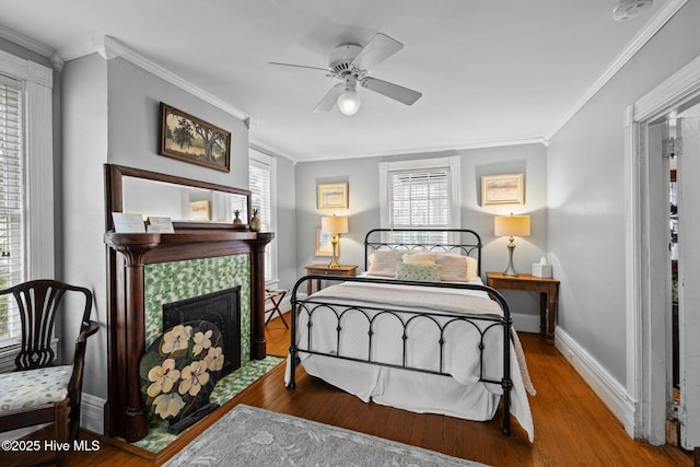 bedroom with a tiled fireplace, wood finished floors, crown molding, baseboards, and ceiling fan