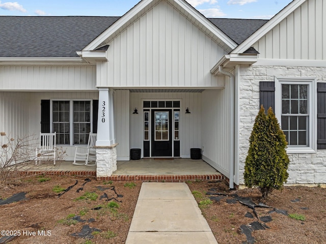 doorway to property with stone siding, a porch, and roof with shingles