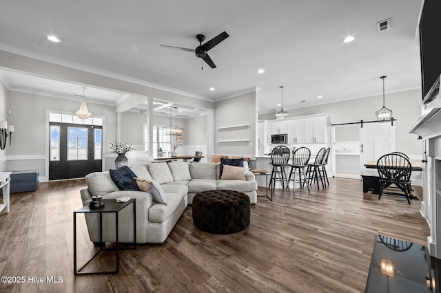 living room featuring a barn door, dark wood-style flooring, visible vents, a ceiling fan, and crown molding