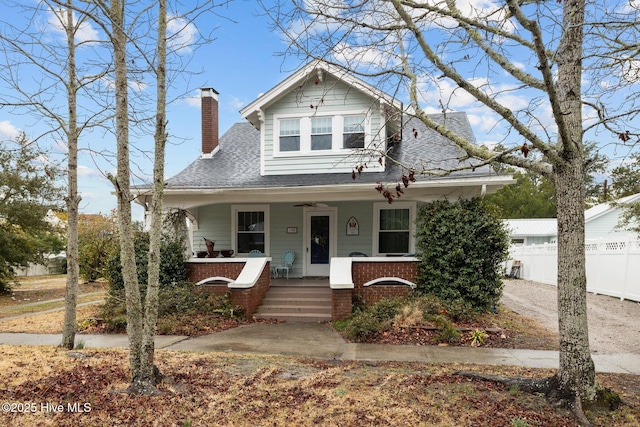 bungalow-style house with a porch, fence, a shingled roof, brick siding, and a chimney