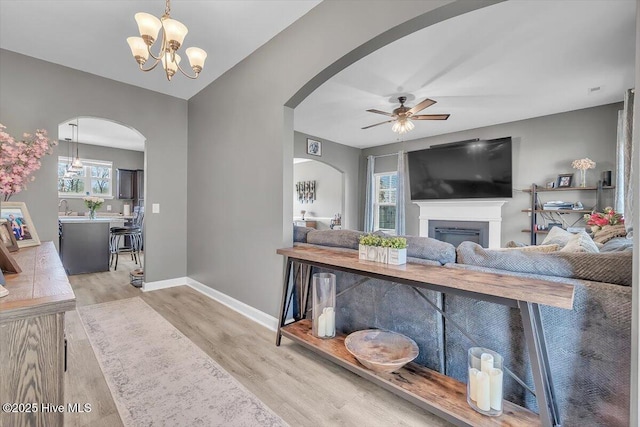 living area featuring ceiling fan with notable chandelier, a glass covered fireplace, wood finished floors, arched walkways, and baseboards