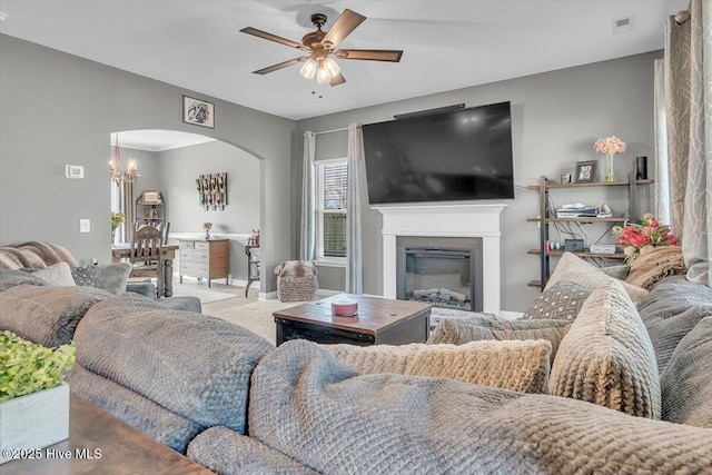 living area featuring carpet, visible vents, arched walkways, a glass covered fireplace, and ceiling fan with notable chandelier