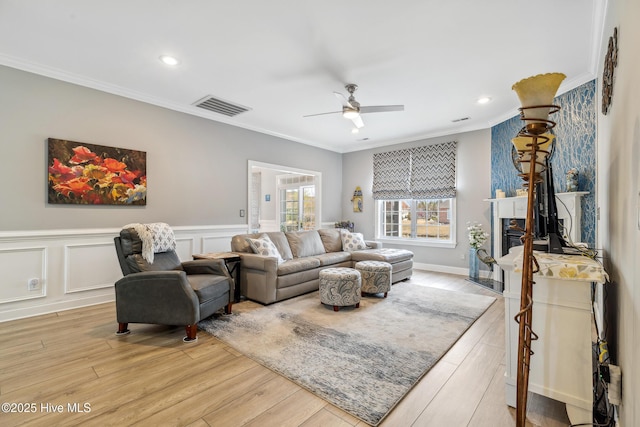 living area featuring a ceiling fan, visible vents, light wood-style flooring, ornamental molding, and a decorative wall