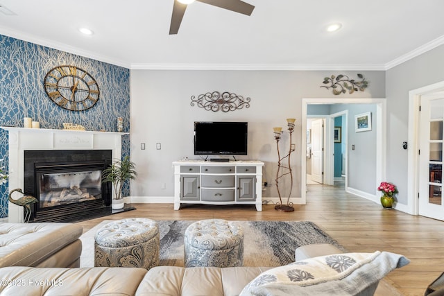 living area with wood finished floors, baseboards, ceiling fan, a glass covered fireplace, and crown molding