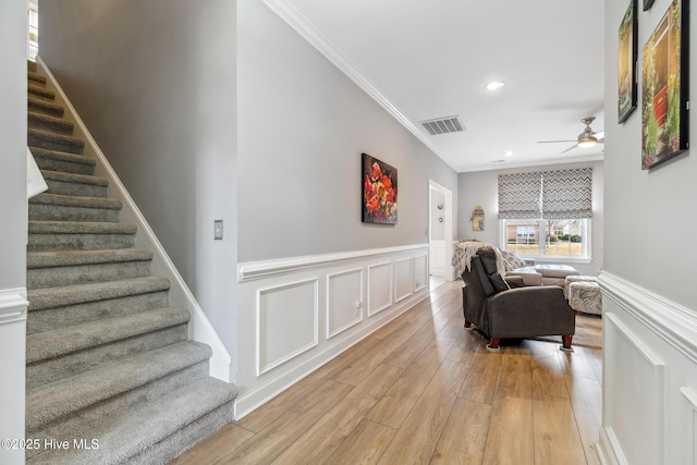 living area featuring stairway, a ceiling fan, visible vents, light wood finished floors, and crown molding