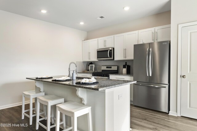 kitchen with wood finished floors, a center island with sink, appliances with stainless steel finishes, white cabinetry, and a kitchen breakfast bar