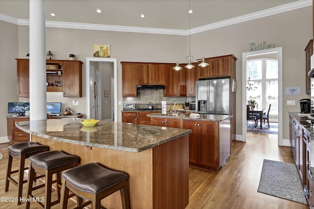 kitchen with light wood finished floors, stainless steel fridge with ice dispenser, a kitchen island with sink, under cabinet range hood, and brown cabinets