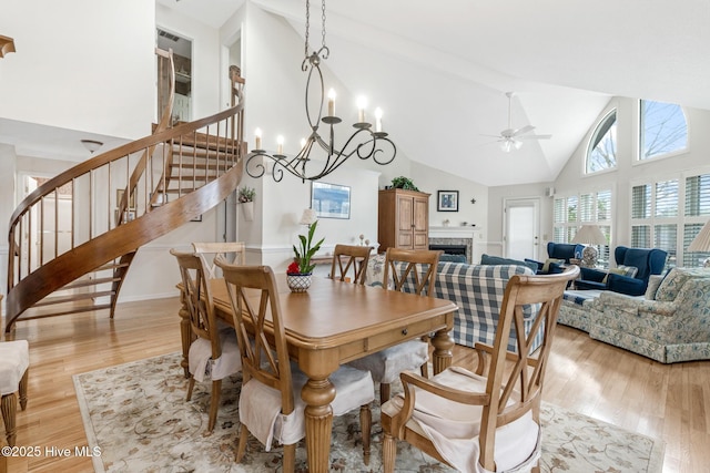 dining room with stairway, high vaulted ceiling, a fireplace, light wood-style floors, and ceiling fan with notable chandelier