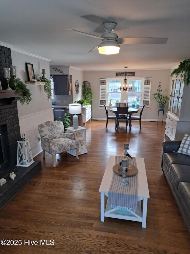 living area featuring a brick fireplace, dark wood-style floors, crown molding, and ceiling fan with notable chandelier