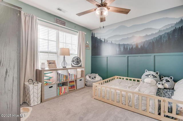 carpeted bedroom featuring visible vents, wainscoting, a decorative wall, and ceiling fan