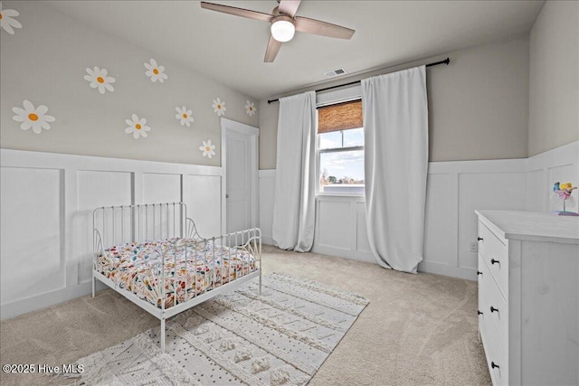 bedroom featuring light carpet, a ceiling fan, wainscoting, and a decorative wall