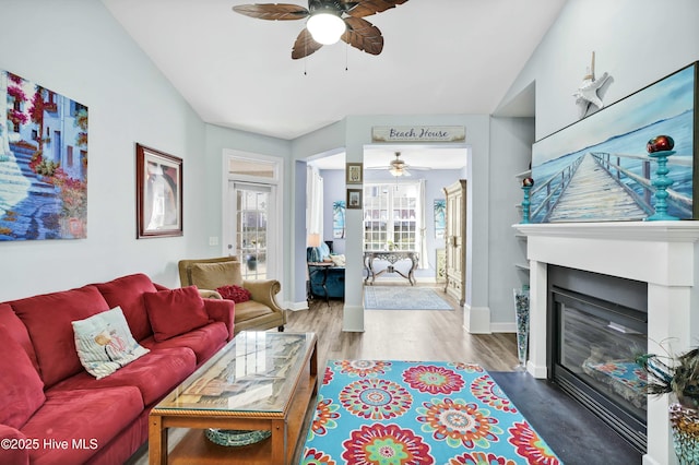 living area with a ceiling fan, a glass covered fireplace, wood finished floors, baseboards, and vaulted ceiling