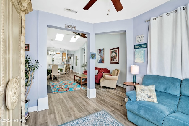 living area with visible vents, baseboards, vaulted ceiling with skylight, light wood-style flooring, and a ceiling fan