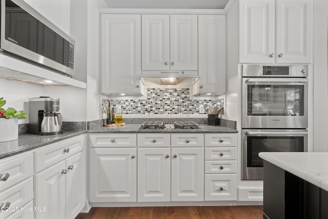 kitchen featuring tasteful backsplash, under cabinet range hood, dark stone counters, appliances with stainless steel finishes, and white cabinets