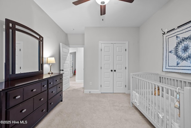 bedroom with a crib, baseboards, light colored carpet, a closet, and a ceiling fan