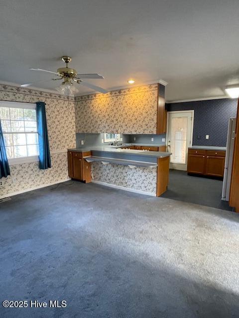 kitchen featuring wallpapered walls, brown cabinetry, dark colored carpet, and ceiling fan