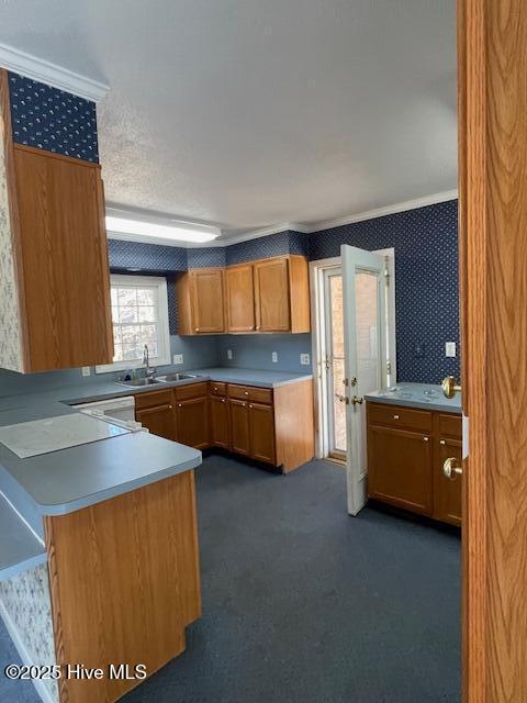 kitchen with ornamental molding, a sink, dark carpet, brown cabinetry, and wallpapered walls