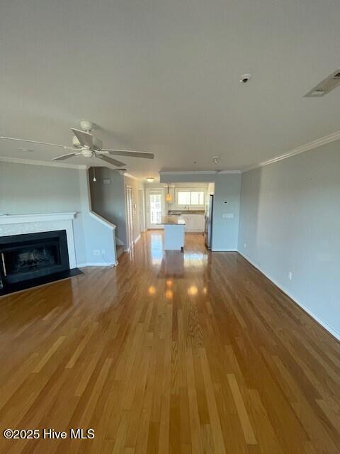 unfurnished living room featuring wood finished floors, visible vents, a fireplace with flush hearth, ceiling fan, and crown molding