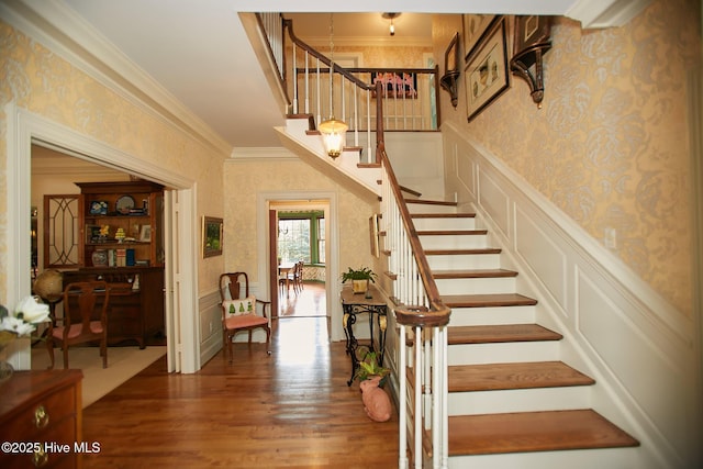 stairway with wood finished floors, wallpapered walls, ornamental molding, wainscoting, and a decorative wall