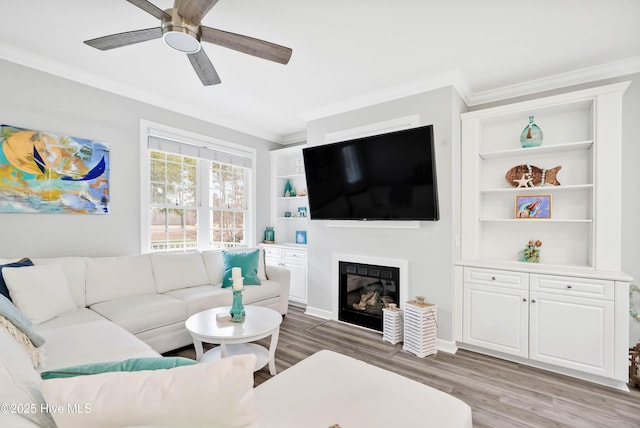 living room with built in shelves, light wood-style flooring, ornamental molding, a glass covered fireplace, and ceiling fan