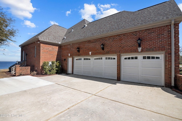 view of side of property featuring a water view, concrete driveway, an attached garage, a shingled roof, and brick siding