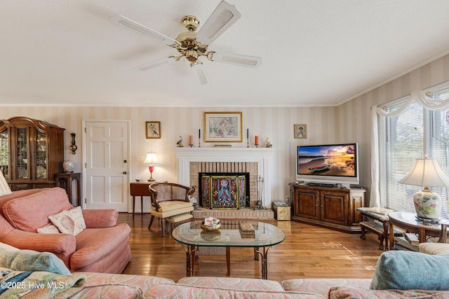 living area featuring wood finished floors, crown molding, wallpapered walls, a brick fireplace, and ceiling fan