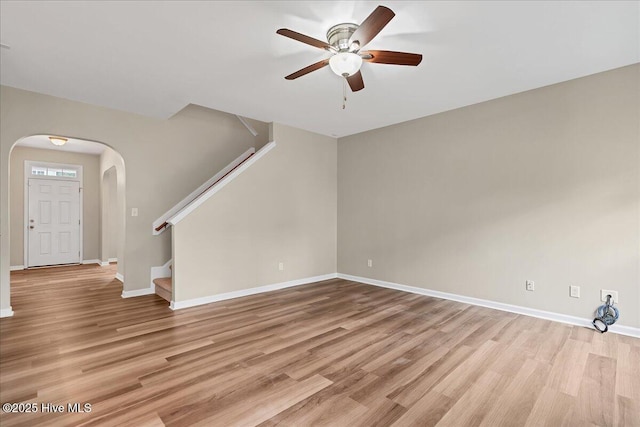 unfurnished living room featuring a ceiling fan, arched walkways, light wood-style floors, baseboards, and stairs