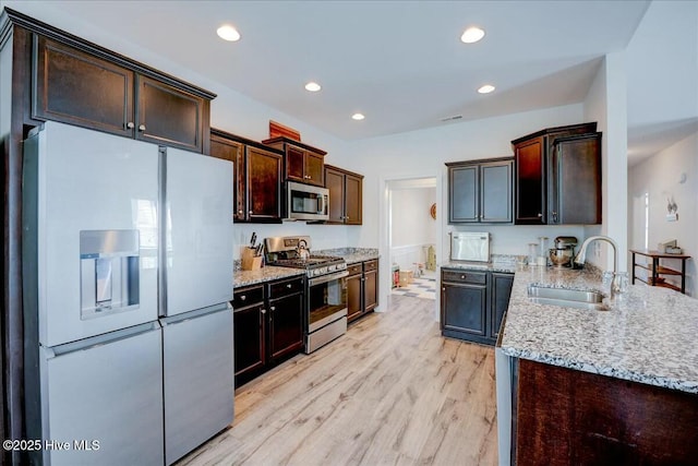 kitchen with a sink, light stone counters, light wood-style floors, appliances with stainless steel finishes, and dark brown cabinets