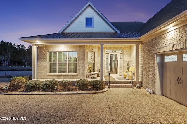 exterior entry at dusk with a garage, brick siding, a porch, and a standing seam roof