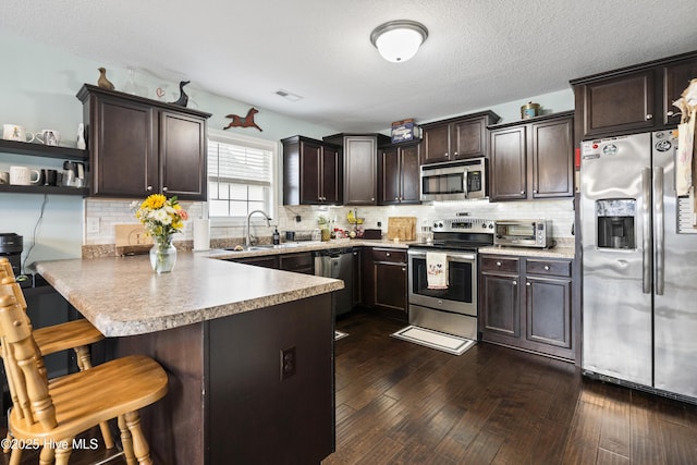 kitchen featuring dark brown cabinets, dark wood-type flooring, a peninsula, stainless steel appliances, and a sink