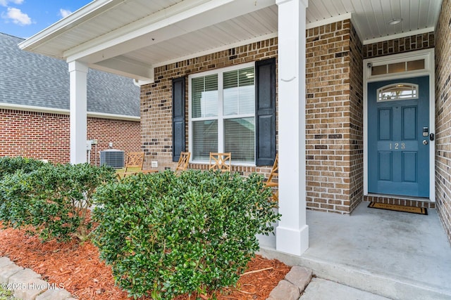 view of exterior entry with covered porch, brick siding, roof with shingles, and central air condition unit
